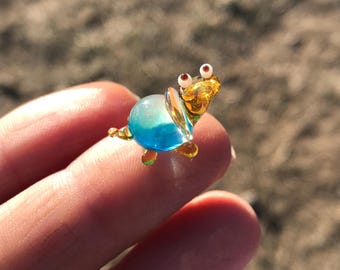 以下が含まれることがあります： 青い殻と黄色の殻、茶色の目をした小さなガラス製の亀の置物。亀は指の上に座っています。