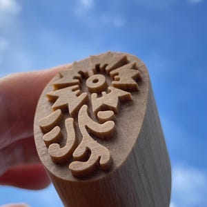 May include: A wooden stamp with a floral design. The stamp has a cylindrical handle and a detailed pattern of stylized leaves and a central circular element. The stamp is a light brown color.