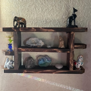 May include: A three-tiered, dark-stained wooden shelf displaying various decorative items. The top shelf holds a black cat statue and an elephant figurine. The middle shelf features a geode and a small statue. The bottom shelf has a cat figurine, rocks, and a ceramic horse.