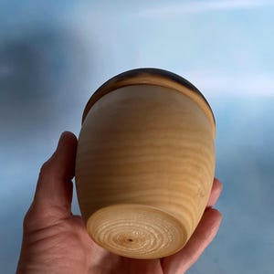 May include: A handcrafted wooden container with a lid. The container is light brown with a darker brown rim on the lid. The wood grain is visible, and the container has a smooth, rounded shape. The container is held in a hand.