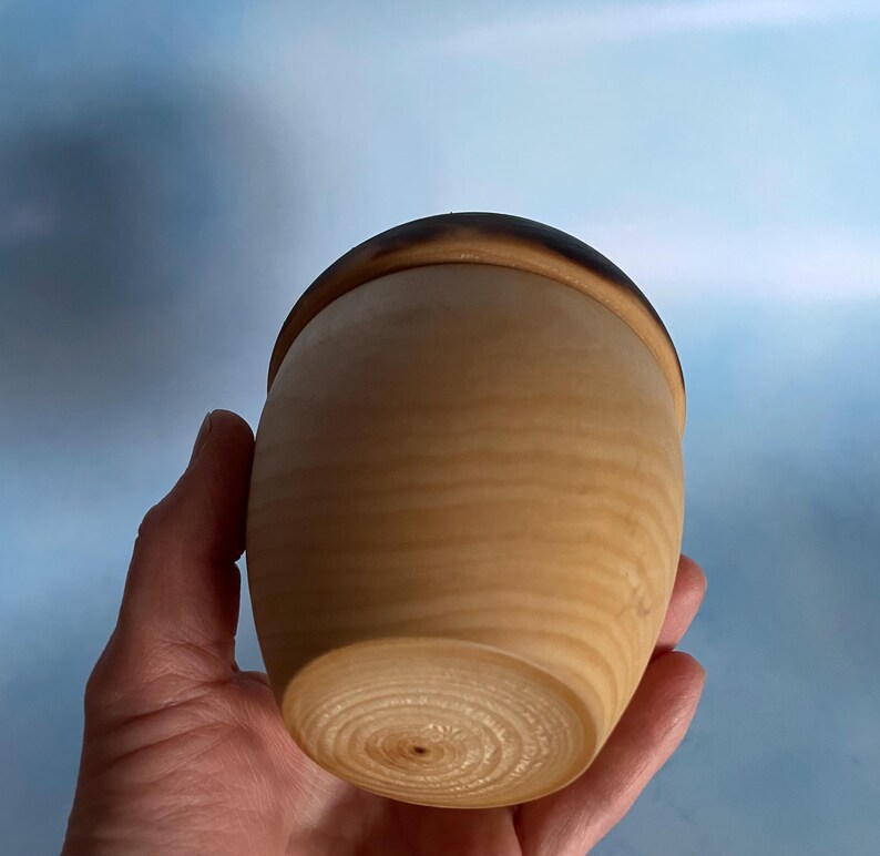 May include: A handcrafted wooden container with a lid. The container is light brown with a darker brown rim on the lid. The wood grain is visible, and the container has a smooth, rounded shape. The container is held in a hand.