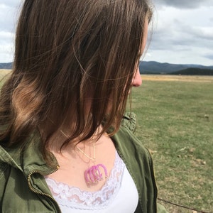 May include: A woman wearing a green jacket and a white tank top with a pink beaded necklace. The necklace features a design of multiple arches.