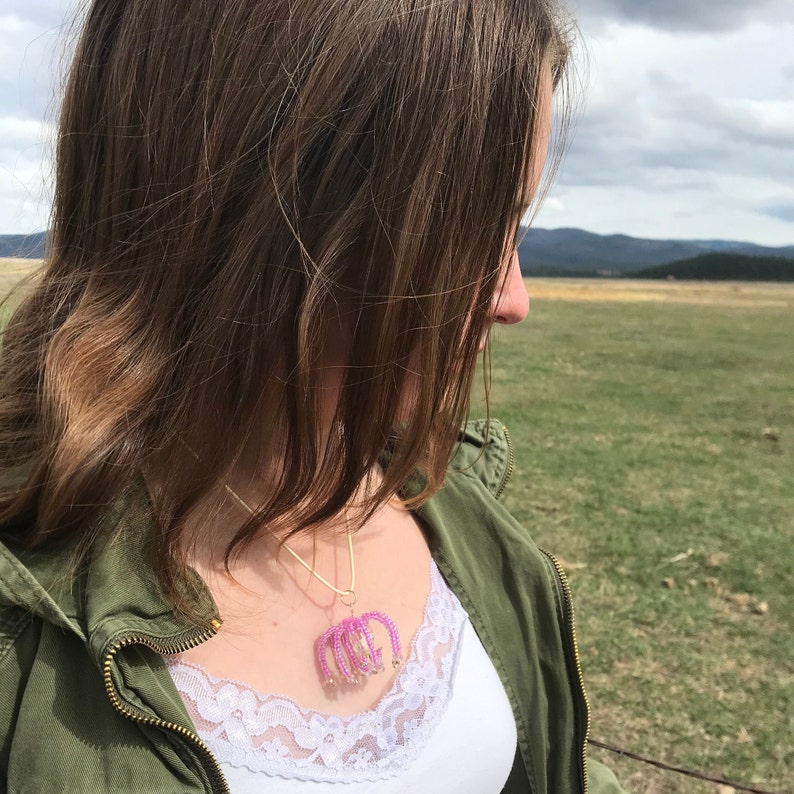 May include: A woman wearing a green jacket and a white tank top with a pink beaded necklace. The necklace features a design of multiple arches.