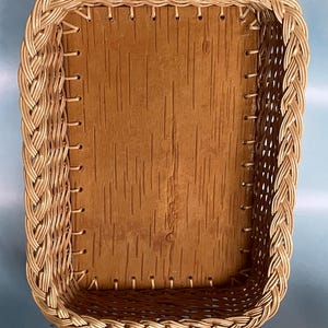 May include: A rectangular basket with a woven, braided border and a light brown, textured interior. The basket's corners are neatly constructed, and the overall design suggests a handcrafted item. The basket is empty and sits on a blue surface.