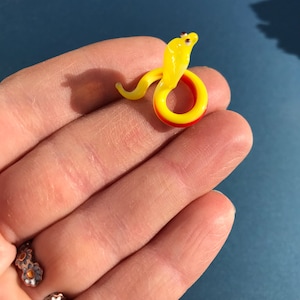 May include: A small glass snake figurine with a yellow body and red and yellow tail. The snake is coiled in a circle and has a raised head.