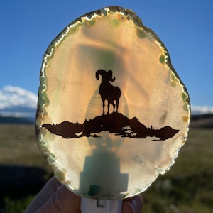 May include: A white agate night light with a silhouette of a bighorn sheep on a mountain. The agate is translucent and has green and brown markings.