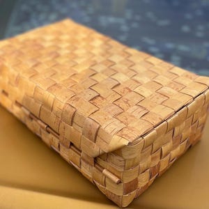 May include: A woven basket made of natural materials, with a light brown color and a rectangular shape. The basket is closed and has a woven lid.