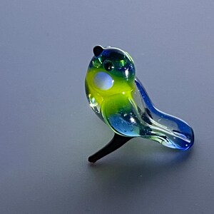 May include: A colorful glass bird figurine. The bird features a combination of green, yellow, and blue hues. It has a black beak and feet. The figurine is a decorative item, likely for display.