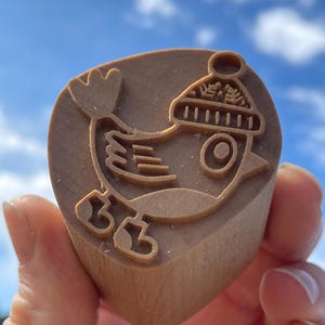 May include: A wooden stamp featuring a bird design wearing a winter hat. The bird has a round body, a large eye, and detailed wing and foot outlines. The stamp is a light brown color.