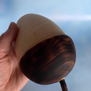 May include: A handcrafted wooden acorn with a light brown body and a dark brown cap. The acorn has a smooth, polished finish and a small stem. The natural wood grain is visible, adding to its rustic charm. The acorn is held against a light blue background.