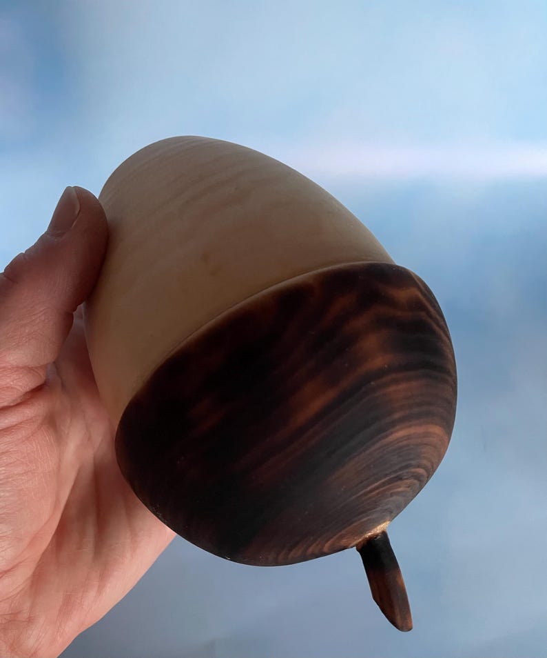 May include: A handcrafted wooden acorn with a light brown body and a dark brown cap. The acorn has a smooth, polished finish and a small stem. The natural wood grain is visible, adding to its rustic charm. The acorn is held against a light blue background.