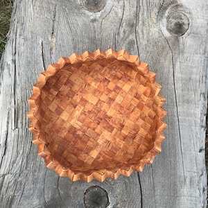 May include: A round, woven basket made from light brown birch bark. The basket has a scalloped edge and a textured, checkered pattern. The basket is displayed on a weathered, gray wooden surface.