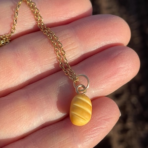 May include: A gold-colored necklace with a small, oval pendant. The pendant is a light yellow color with diagonal carved lines. The necklace chain is delicate and appears to be gold-plated. The necklace is shown against a blurred background.