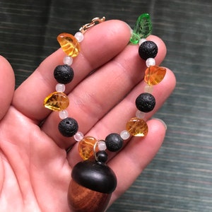 May include: A handmade bracelet featuring a wooden acorn pendant with a dark brown cap and a light brown base. The bracelet includes amber-colored glass beads, black lava beads, and small white beads, connected by a gold-colored chain.