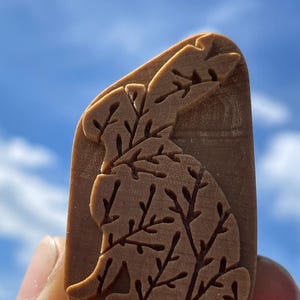 May include: A brown rectangular stamp with a detailed relief of a rabbit and branches. The stamp is held against a blue sky background. The rabbit design is a light brown color.