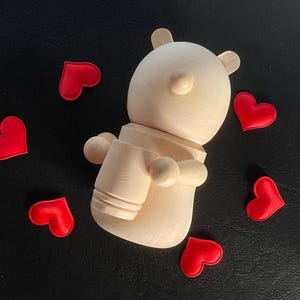 May include: A wooden bear-shaped figurine with a light brown finish, featuring rounded ears and arms. Scattered around the bear are several small, bright red heart-shaped embellishments. The objects are set against a dark background.