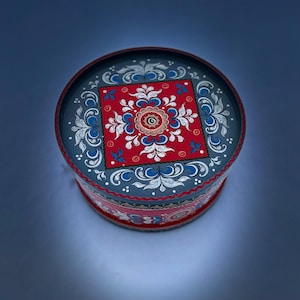May include: A round, gray painted wooden box with a red and blue floral design. The box has a lid and is decorated with traditional Scandinavian folk art.