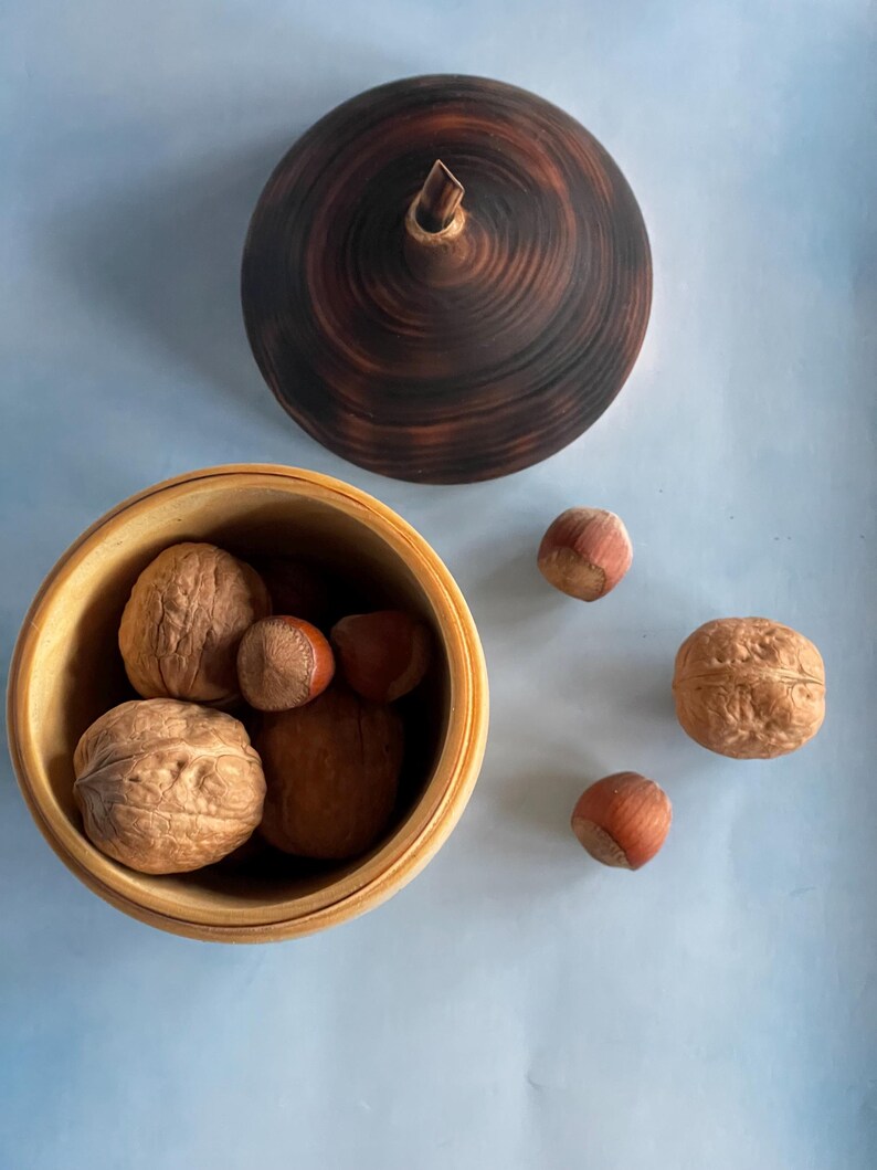 May include: A wooden bowl filled with walnuts and hazelnuts, with a matching lid. The lid and bowl are a light brown color. Several walnuts and hazelnuts are scattered around the bowl on a light blue surface.