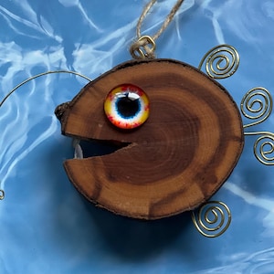 May include: A wooden fish ornament with a large, colorful eye and a wire lure. The fish is brown with a natural wood grain and has a wire tail with three curls.