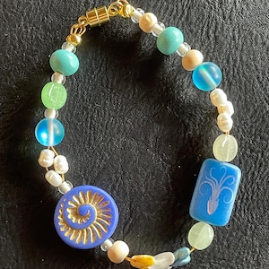 May include: A beaded bracelet featuring a variety of colorful beads. The bracelet includes blue, green, and cream-colored beads, along with a blue circular bead with a gold spiral design and a blue rectangular bead with a white octopus design.