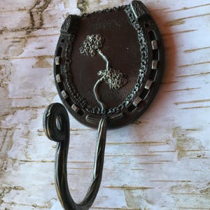 May include: A black metal horseshoe hook with a decorative tree design. The hook is attached to a wall.