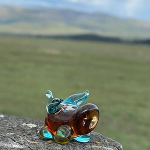 May include: A small, glass snail figurine with a brown body, blue wings, and yellow accents. The snail is perched on a weathered wooden post, with a blurred green and blue landscape in the background.