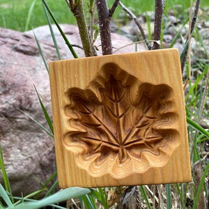 May include: A square, wooden mold with a carved maple leaf design. The mold is made of light-colored wood and features detailed leaf veins. The background includes green grass and a blurred natural setting.