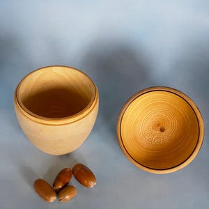 May include: A wooden, acorn-shaped container with a lid, crafted from light-colored wood. The lid is off, revealing the interior. Four acorns are placed near the container, suggesting a natural theme.