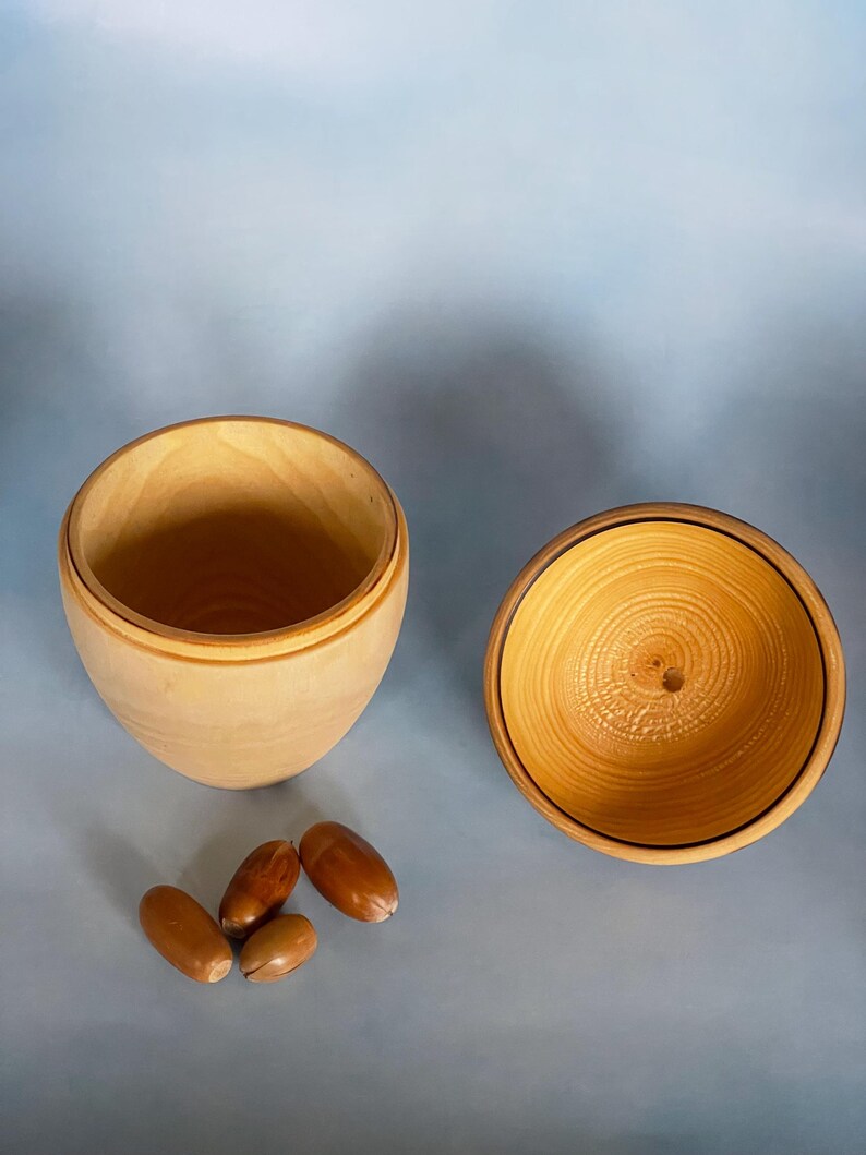 May include: A wooden, acorn-shaped container with a lid, crafted from light-colored wood. The lid is off, revealing the interior. Four acorns are placed near the container, suggesting a natural theme.