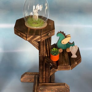 May include: A tiered, wooden display stand with a dark stain. The top shelf holds a glass egg with a bunny figurine inside. The middle shelf features a wooden dragon toy, a small succulent in an orange pot, and a carved angel figurine.