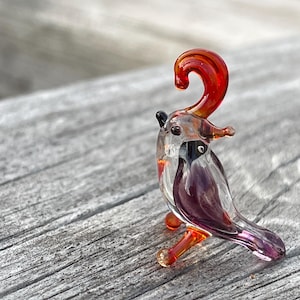 May include: A small glass bird figurine with a red and orange crest, purple body, and orange legs. The bird is perched on a wooden surface.