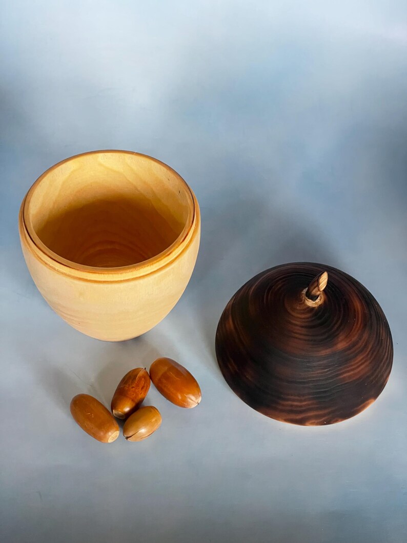 May include: A wooden acorn-shaped box with a dark brown lid and a light-colored interior. The lid has a small stem. Four acorns are next to the box. The box is open, revealing the interior.