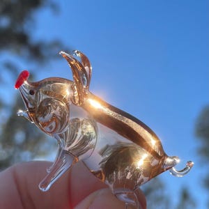 May include: A small, clear glass pig figurine with gold and brown accents. The pig has a red nose and is held in a person's hand against a blue sky background. The figurine is detailed and reflects light.