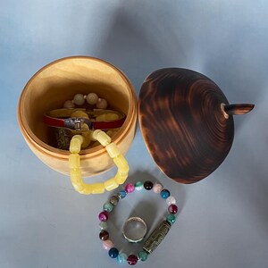May include: A wooden jewelry box with a dark brown lid and a light wood interior. Inside the box are several bracelets and a red bracelet. A beaded bracelet and a silver ring are next to the box. The background is a light blue.