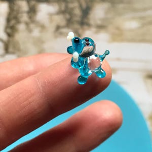 May include: A small, blue glass figurine of a dog holding a bone in its mouth. The figurine is made of clear glass with blue accents and is about 1 inch tall.