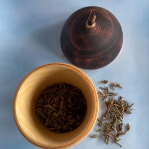 May include: A wooden bowl filled with dried tea leaves next to a small pile of loose leaves. A dark brown, wooden lid with a pointed top sits nearby. The bowl and lid are on a light blue surface.