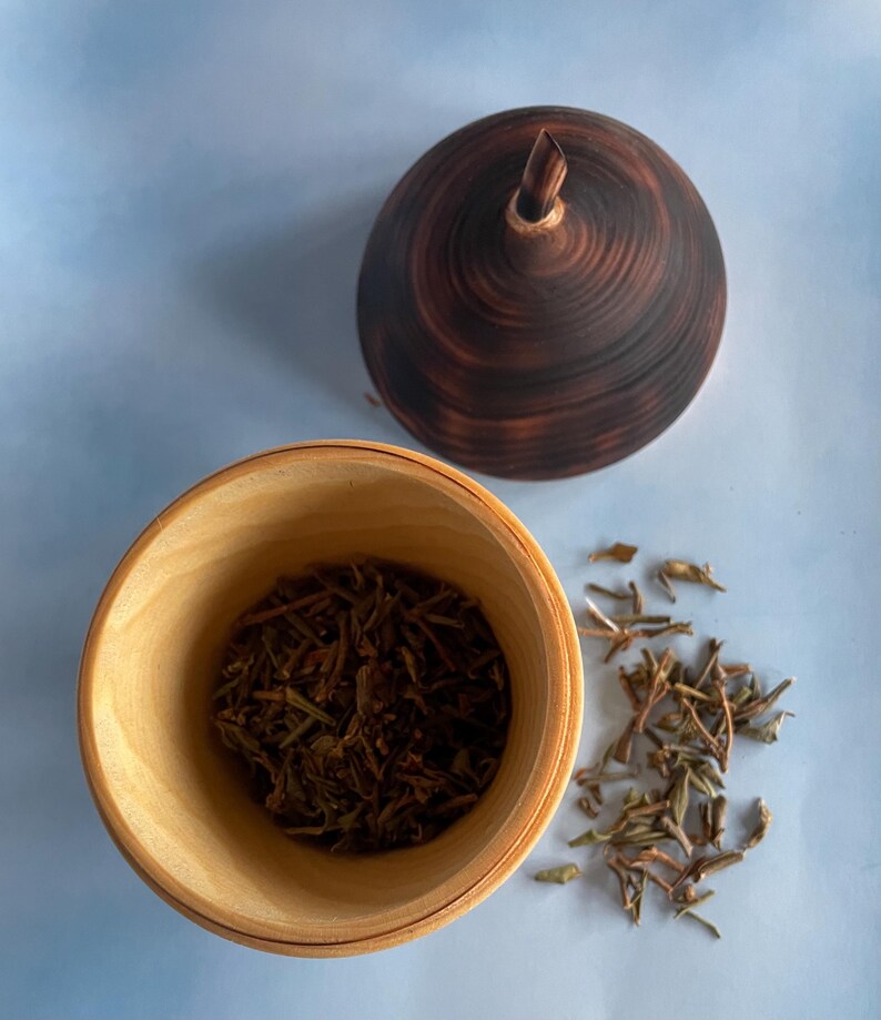 May include: A wooden bowl filled with dried tea leaves next to a small pile of loose leaves. A dark brown, wooden lid with a pointed top sits nearby. The bowl and lid are on a light blue surface.