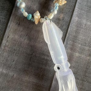 May include: A handmade bracelet featuring a clear, iridescent squid charm. The bracelet includes seashells, pearls, and beads in shades of white, silver, and turquoise. The bracelet is secured with a silver clasp.