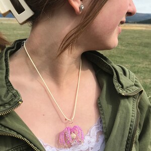 May include: A close-up of a person wearing a green jacket and a necklace with a pink beaded pendant. The pendant is shaped like a flower with a white center.