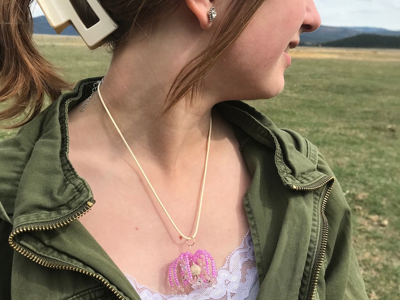 May include: A close-up of a person wearing a green jacket and a necklace with a pink beaded pendant. The pendant is shaped like a flower with a white center.