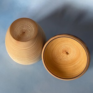May include: Two wooden bowls, one with the lid off, showcasing the natural wood grain. The bowls are light brown with a darker rim on the open bowl. The wood grain is visible on the inside and outside of the bowls.