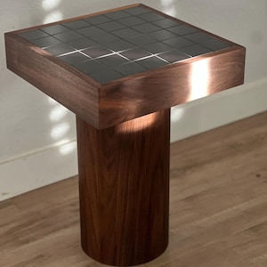 May include: A modern side table with a square top and a cylindrical base. The top is inlaid with a grid of dark, reflective tiles, framed by a rich, dark wood. The base is made of the same wood, creating a cohesive design.