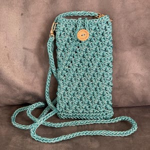 Op de afbeelding: Een teal gehaakte schoudertas met een gestructureerd steekpatroon, een lange gevlochten riem met goudkleurige metalen sluitingen en een ronde houten knoopsluiting gegraveerd met de tekst Handmade with love en een klein hartje.