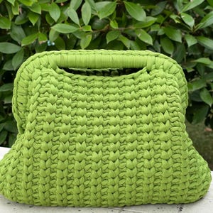 May include: A lime green crocheted handbag with a rectangular shape and a handle. The bag is made of thick yarn, with a textured, woven appearance. The background features green foliage.