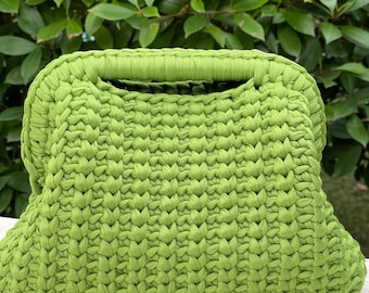 Könnte beinhalten: Eine hellgrüne gehäkelte Handtasche mit rechteckiger Form und Griff. Die Tasche ist aus dickem Garn gefertigt und hat ein strukturiertes, gewebtes Aussehen. Der Hintergrund zeigt grünes Laub.