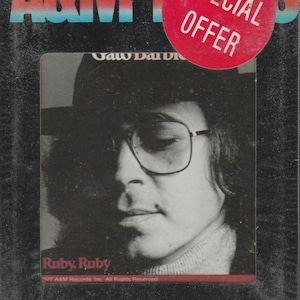 May include: A vintage cassette tape case featuring a black and white portrait of a person wearing glasses and a hat. The text "Ruby Ruby" is visible, along with "A&M Records" and a "Special Offer" sticker. The case is from 1977.