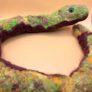 May include: A handmade felt snake in shades of green, yellow, and purple. The snake has a detailed head with a green eye and a curved body. The felted texture gives the snake a soft, plush appearance, suitable for a child's toy or decorative item.