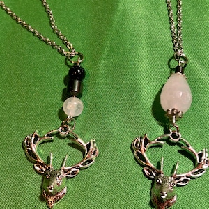May include: Two silver necklaces featuring stag head pendants. One has black and white beads, the other a pink, teardrop-shaped bead. The pendants have detailed antlers and a textured finish, set against a green backdrop.