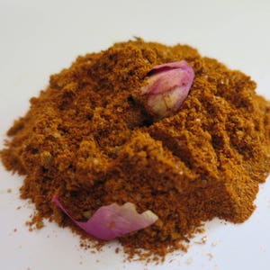 May include: A mound of finely ground, rust-colored spice blend featuring a single dried pink rosebud nestled near the top and a loose pink rose petal resting at the base of the pile, showcasing a textured, granular consistency throughout.