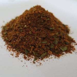 May include: A pile of reddish-brown spice blend with visible green herbs. The spices are finely ground and appear to be a mix of different herbs and spices. The image is taken on a white surface.